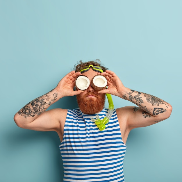 Free Photo funny playful bearded man wears snorkel mask, prepares for diving underwater, keeps two coconuts on eyes