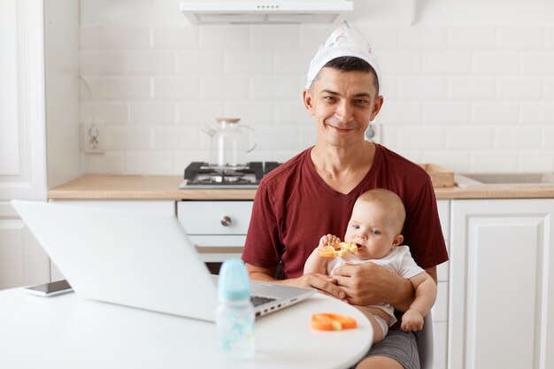 Funny positive attractive brunette man freelancer wearing casual style maroon t shirt and diaper on his head, working and looking after his little baby daughter.