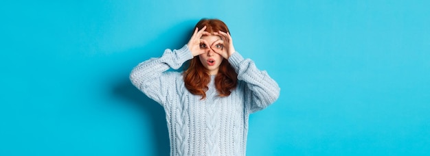 Free Photo funny redhead female model in sweater staring at camera through fingers glasses seeing something int