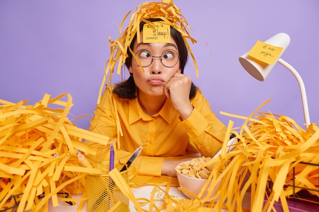 Free Photo funny schoolgirl prepares for test at home has sleepless night works late hours makes grimace has busy working schedule surrounded by paper wastes isolated over purple wall