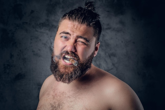 Funny shirtless bearded male smoking a cigar on grey background.