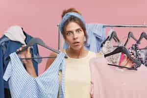 Free photo funny shocked young woman wearing headband holding two hangers with fashionable clothing items in each hand having a desire to buy them both while shopping in store in big sale. consumerism concept