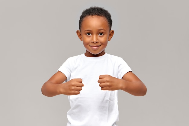 Free photo funny ten year old african boy in white t-shirt keeping clenched fists in front of him demonstrating strength or holding invisible objects