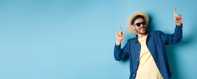 Free photo funny tourist dancing on vacation pointing fingers up wearing summer hat and sunglasses blue backgro