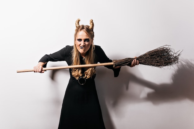 Free Photo funny witch with blonde hair standing on white wall. joyful vampire posing with broom in halloween.