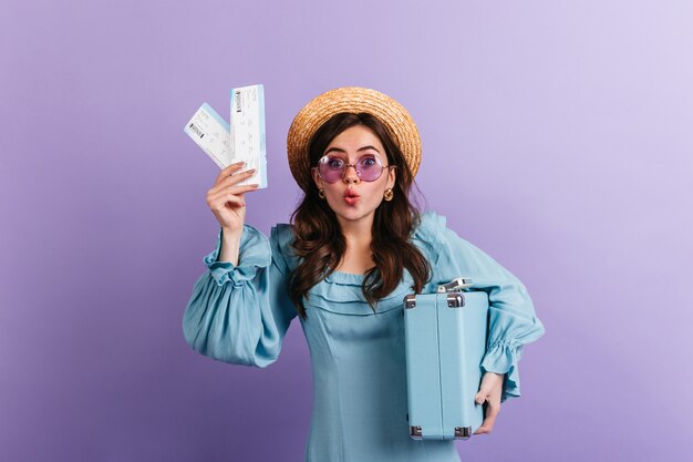 Funny woman in boater and lilac glasses stares in amazement, showing her plane tickets and blue retro suitcase.