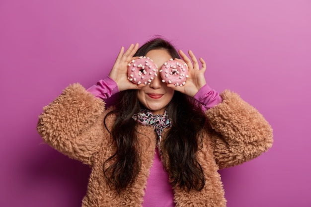 Free photo funny woman covers eyes with decorated doughnuts as glasses, smiles pleasantly, dressed in winter clothing, has fun with desserts