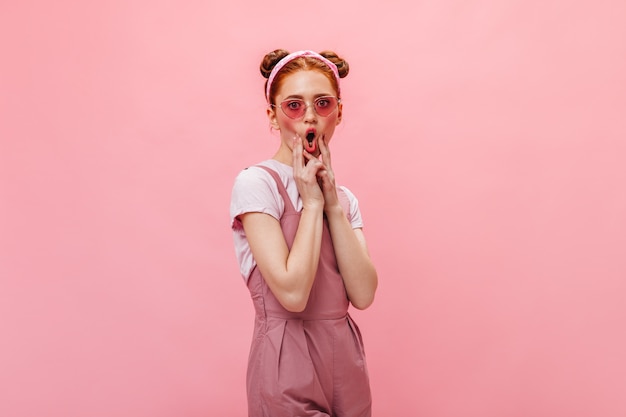 Funny woman with buns makes face. Woman in pink outfit and glasses posing on isolated background.