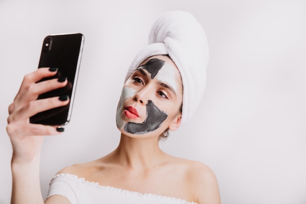 Funny woman with clay mask for face care and in towel on her head makes selfie on white wall.