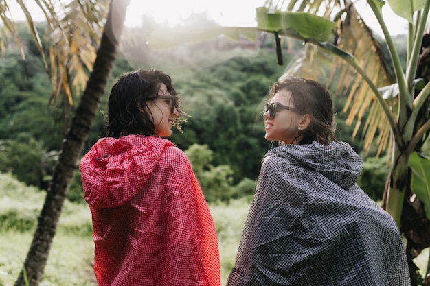 Free photo funny women with wet hair looking at each other while walking around forest. outdoor photo of female tourists in raincoats standing on nature.