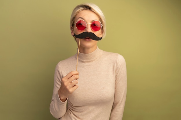 Free photo funny young blonde woman wearing sunglasses holding fake mustache on stick above lips looking at front isolated on olive green wall with copy space