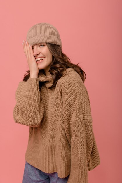 Funny young caucasian brunette girl in hat covers half face with hand on pink background People emotions lifestyle and fashion concept