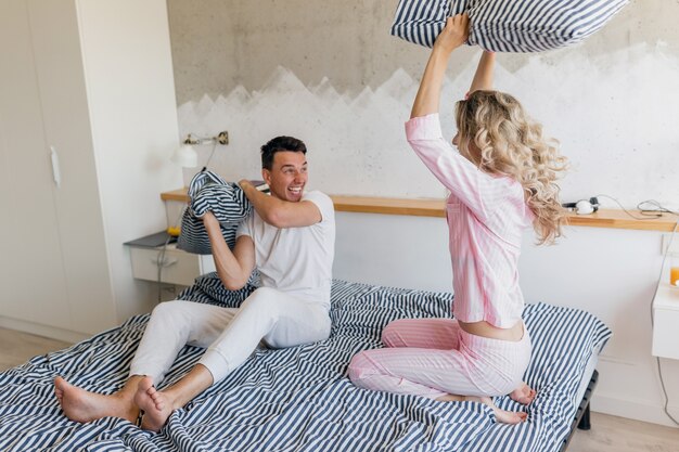 Funny young couple having fun on bed in morning, fighting with pillows, playing, smiling happy