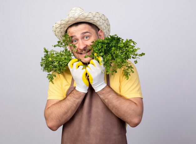 Funny young gardener man wearing jumpsuit and hat in working gloves holding fresh herbs looking at front smiling with happy face standing over white wall