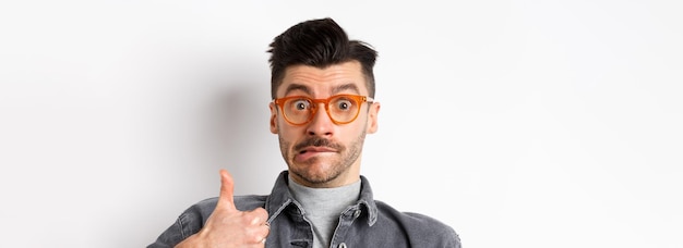 Funny young man in glasses show thumb up biting lip and look indecisive standing on white background