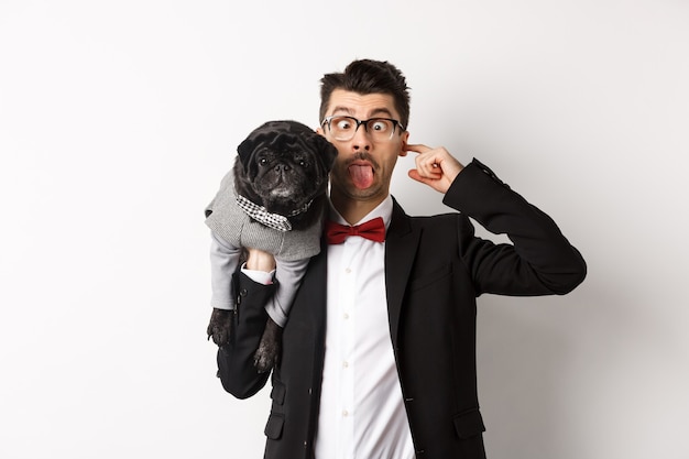 Free Photo funny young man in party suit, showing tongue and holding cute black pug on shoulder, celebrating with pet, standing over white background.