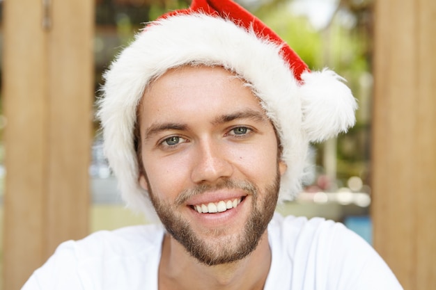 Free photo funny young man in red hat with white fur smiling happily, enjoying holidays in country with hot climate.