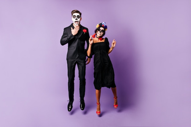 Funny young people with face art on Halloween emotionally pose, jumping on purple background.