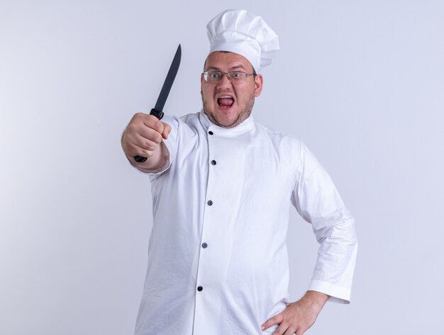 furious adult male cook wearing chef uniform and glasses keeping hand on waist looking at front stretching out knife towards front isolated on white wall