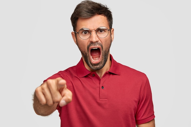 Free photo furious depressed male with dark bristle, yells angrily at someone, points, dressed in bright red tshirt, isolated over white wall. unshaven mad man expresses rage, shouts loudly