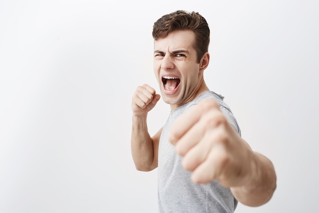 Free photo furious outraged caucasian man shouts in anger, frowns his face, keeps fists, going to defend himself in fight with criminals. desperate european guy dark hair shows his power, clenches fists.