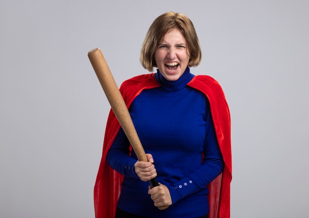 Furious young blonde superhero girl in red cape holding baseball bat  screaming isolated on white wall with copy space