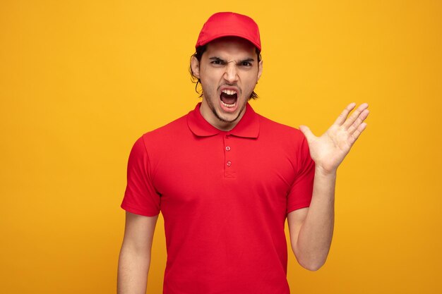 furious young delivery man wearing uniform and cap looking at camera showing empty hand screaming isolated on yellow background