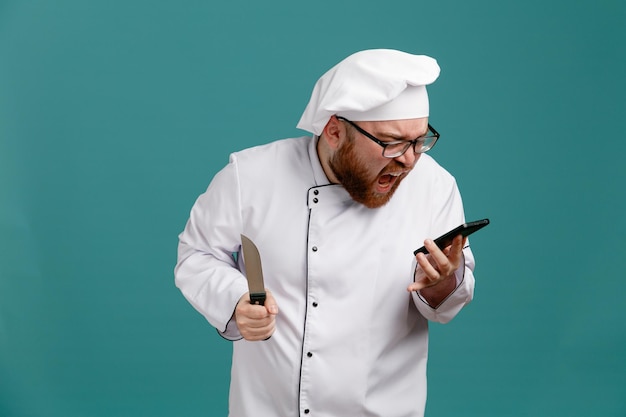 Furious young male chef wearing glasses uniform and cap holding knife holding and looking at mobile phone shouting out loudly isolated on blue background