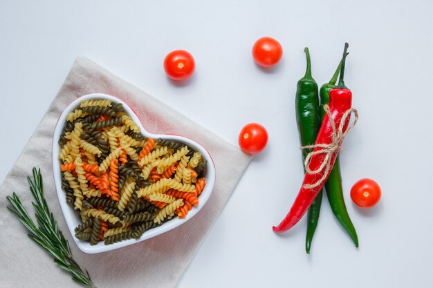 Fusilli pasta in a bowl with peppers, tomatoes, green plant top view on white and folded tablecloth table