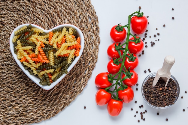 Fusilli pasta in a bowl with tomatoes, peppercorns, scoop top view on white and wicker placemat table