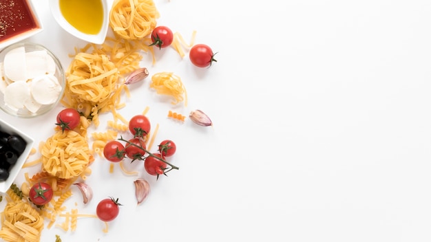 Free Photo fusilli and tagliatelle raw pasta with oil; sauce; olives; cheese; tomato and garlic clove above white surface