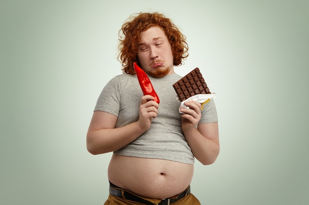 Fustrated funny young redhead man wearing small grey t-shirt with his big belly sticking out, holding red pepper in one hand and bar of chocolate in other doesn't want to eat unappetizing vegetable