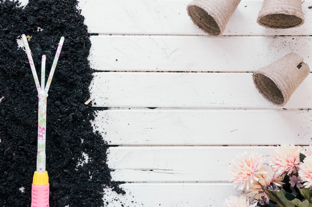 Gardening rake on soil over wooden table with peat pot and flowers