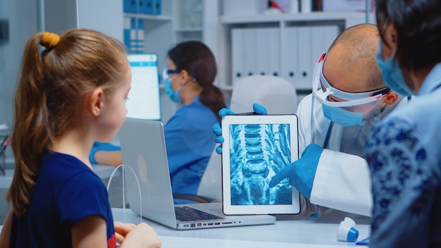 Free photo general practitioner presenting x-ray of neck. pediatrician specialist with protection mask providing health care services, radiographic treatment examination during coronavirus in hospital cabinet