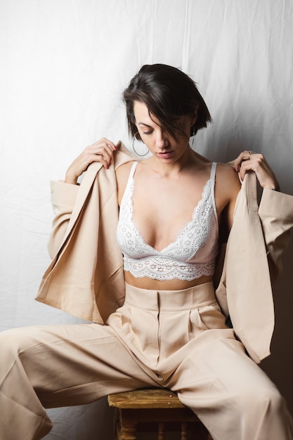 Gentle portrait of a beautiful young woman with big breasts in a beige suit and white lace bra sits on a chair on a gray white