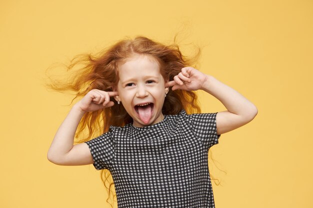 Genuine human emotions, reaction and body language. Angry spoiled little girl with red hair sticking out tongue, pretending not to hear you, plugging ears, screaming, being mad and naughty