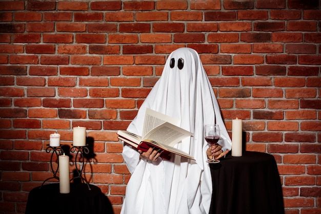 Free photo ghost holding book and wine over brick wall. halloween party.