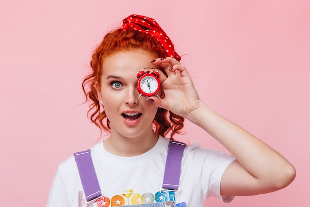 Free photo ginger curly woman with blue eyes looks at front and holds red alarm clock on pink wall