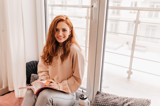 Free Photo ginger emotional girl reading book. romantic stylish lady sitting beside window.