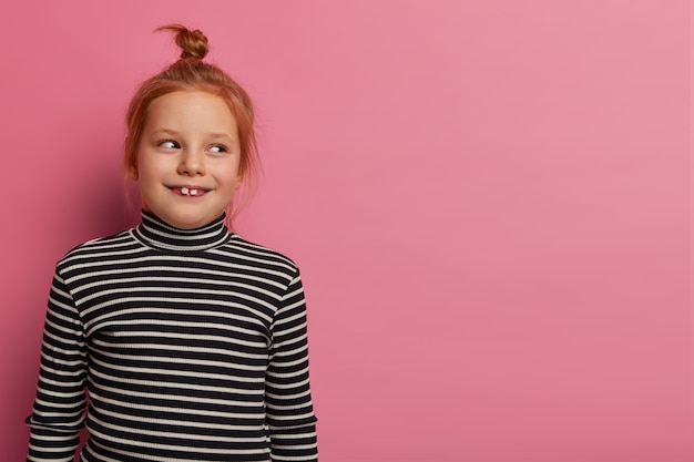 Free photo ginger girl looks gladfully aside, has two teeth stick out, wears striped jumper, being in good mood after coming home from kindergarten, poses indoor over rosy wall. children concept