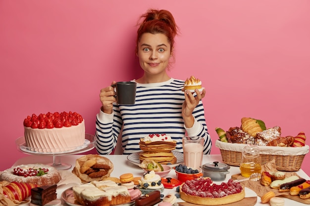 Free photo ginger girl sits at festive table overloaded with many sweet desserts, eats yummy fresh baked cake and drinks tea, has unhealthy but tasty lunch, feels hungry, being voluptuous.