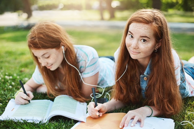 ginger hair gazing with lifted eyebrows and cute smile, lying on grass in city park with sister, sharing earphones to listen music together and doing homework. Lifestyle and people concept