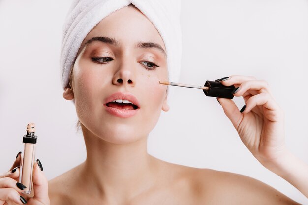 Girl after shower put concealer under eyes. Portrait of beautiful woman on white wall.