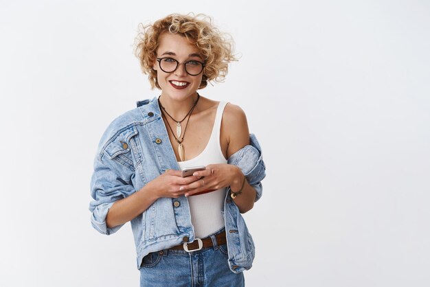 Girl asking number and writing in smartphone smiling broadly at camera friendly posing carefree in relaxed pose in glasses and denim jacket checking grocery list in mobile phone over white background