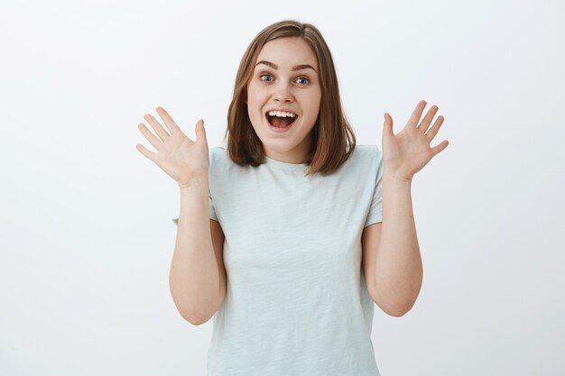 Girl being excited and surprised seeing friend unexpectedly raising palms near shoulder gasping and smiling joyfully from amazement and positive emotions standing in trendy t-shirt over gray wall