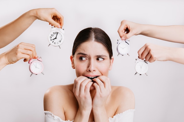 Girl biting her fingers in fright. Woman in fear poses against wall of hands holding clock.