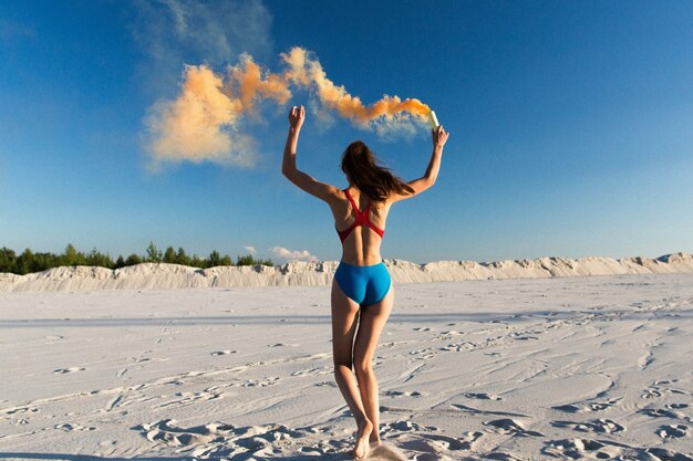 Girl in blue swim-suit dances with orange smoke on white beach