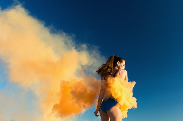 Girl in blue swim-suit dances with orange smoke on white beach