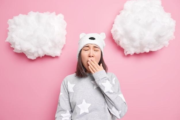 Free photo girl cons mouth yawns wants to sleep has problem of insomnia dressed in nightwear soft bear hat isolated on pink