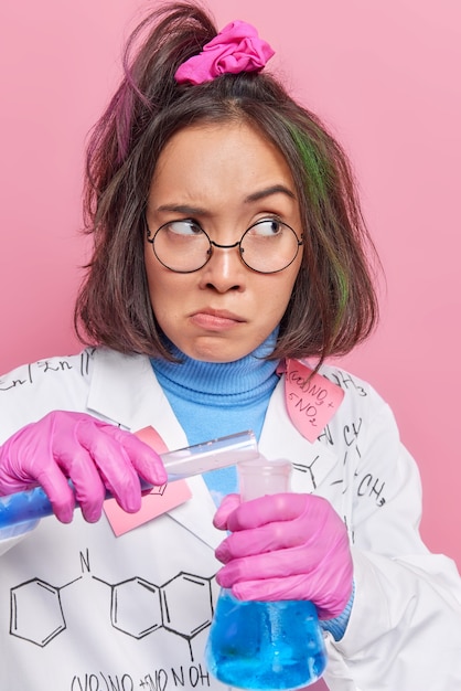 Free photo girl does research with chemical fluid works in laboratory with liquid samples wears round spectacles and medical coat poses on pink